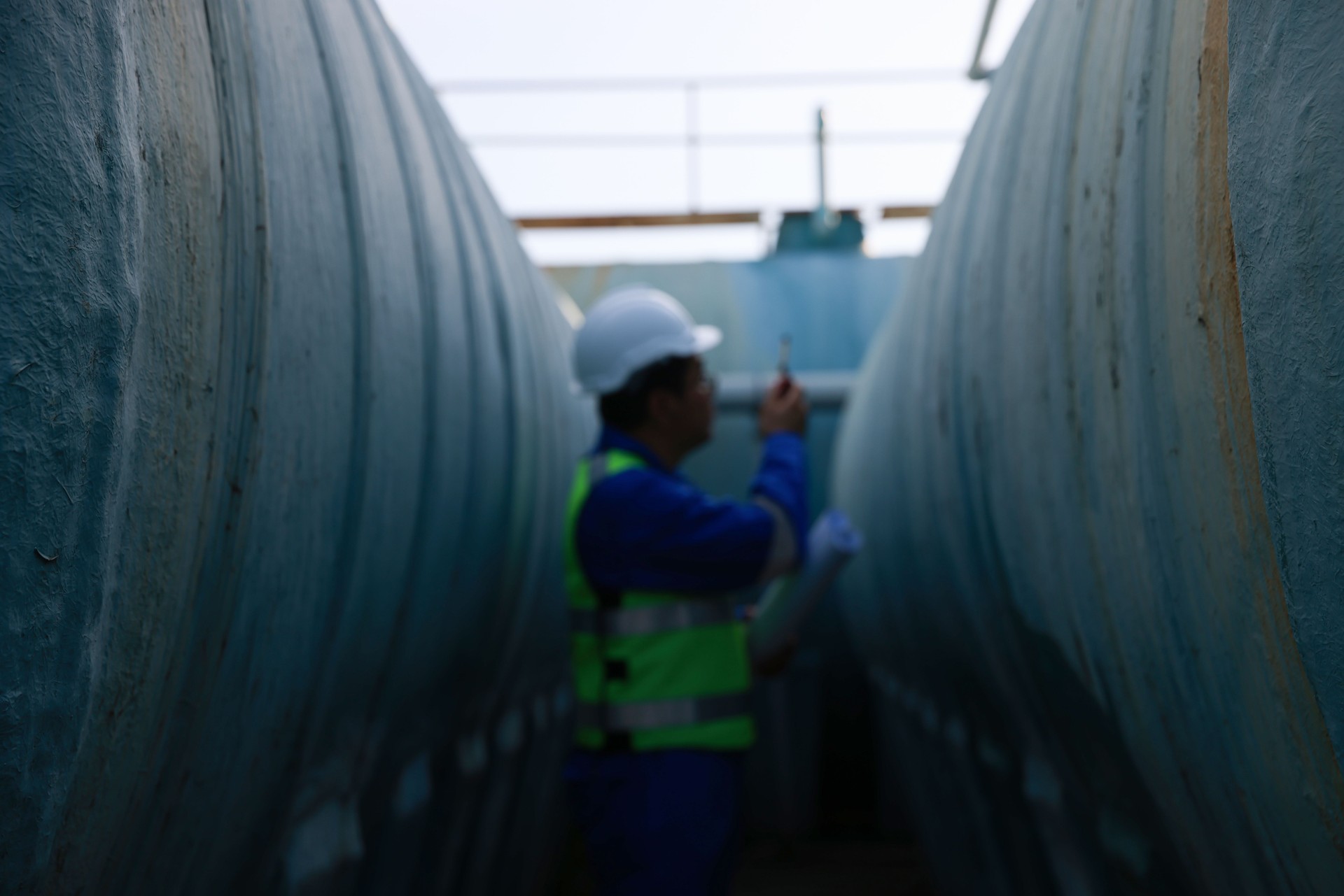 Senior maintenance engineer wearing a reflective vest and using a walkie-talkie while working with a blueprint drawing at a large water tank in a construction site of the factory.